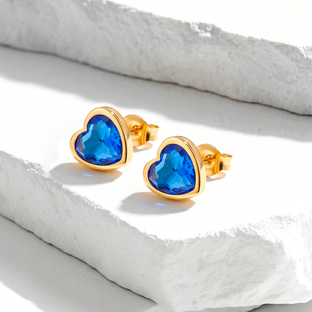 Aretes de broquel para mujer de acero quirúrgico 316 con baño de oro de 18k, con un bisel dorado liso que enmarca un cristal facetado en forma de corazón color azul.