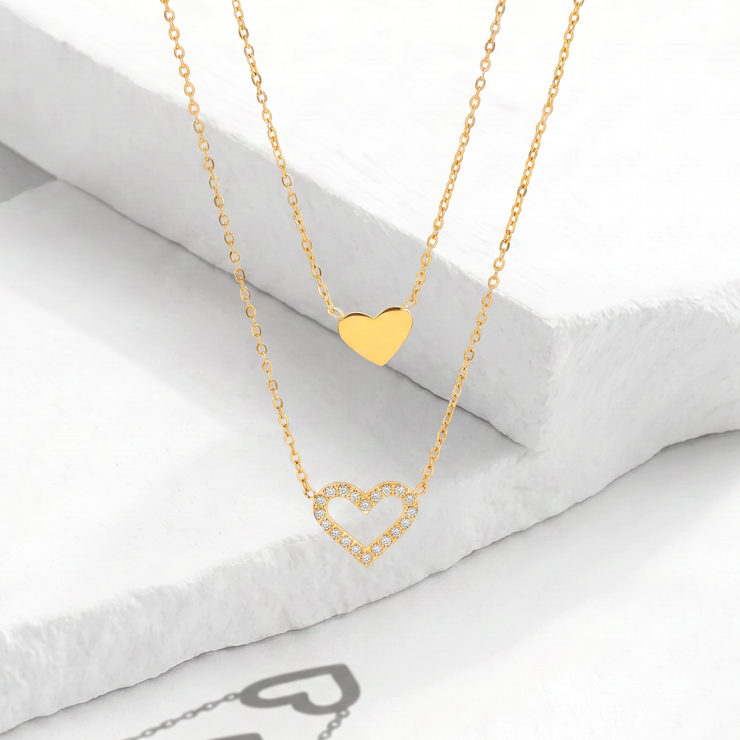Collar de doble capa para mujer marca Bodo en acero inoxidable bañado en oro de 18k con un dije de corazón pulido liso y un dije de corazón calado con cristales blancos.
