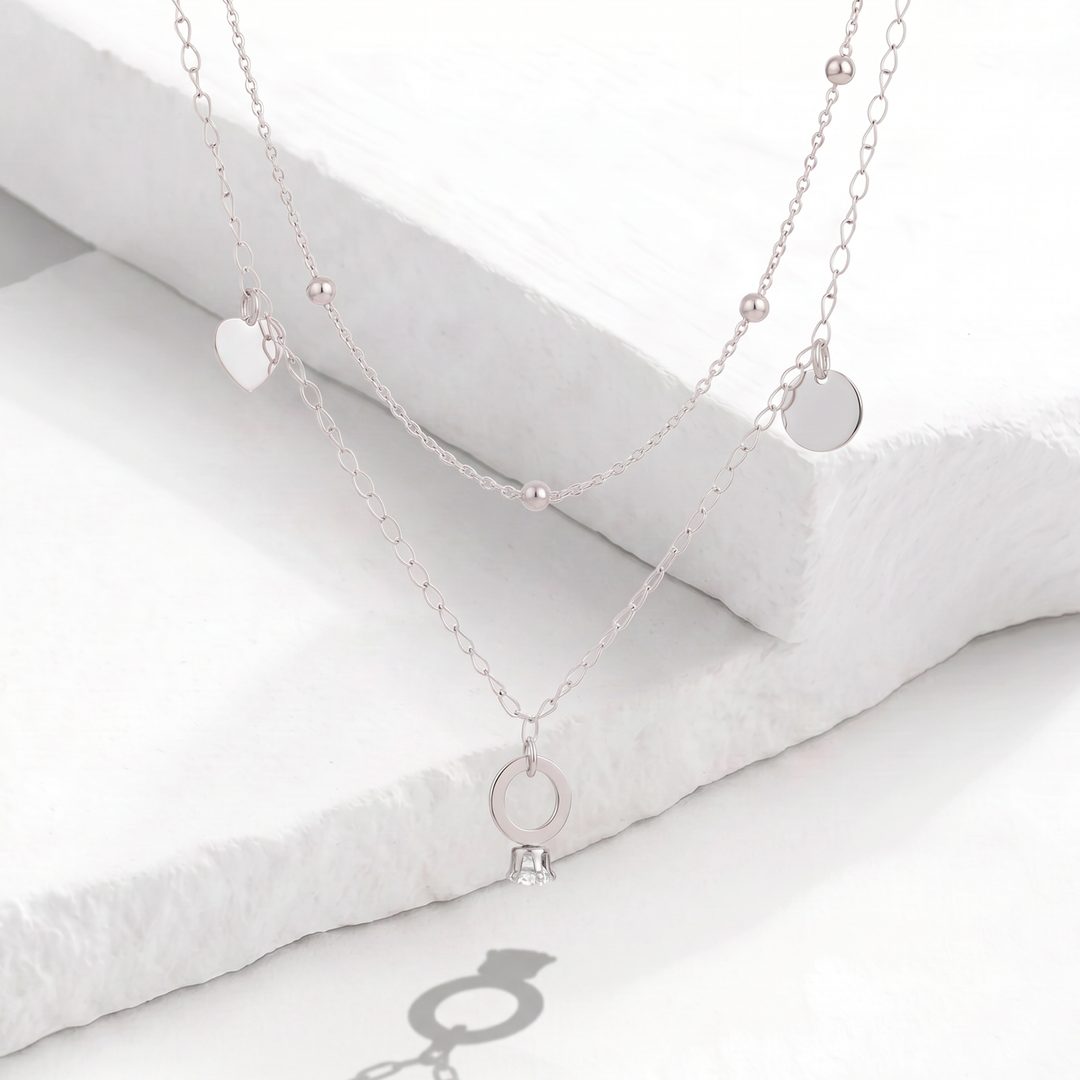 Collar doble cadena para mujer en acero inoxidable plateado con esferas intercaladas y dijes colgantes de corazón, círculo liso y anillo con cristal blanco.