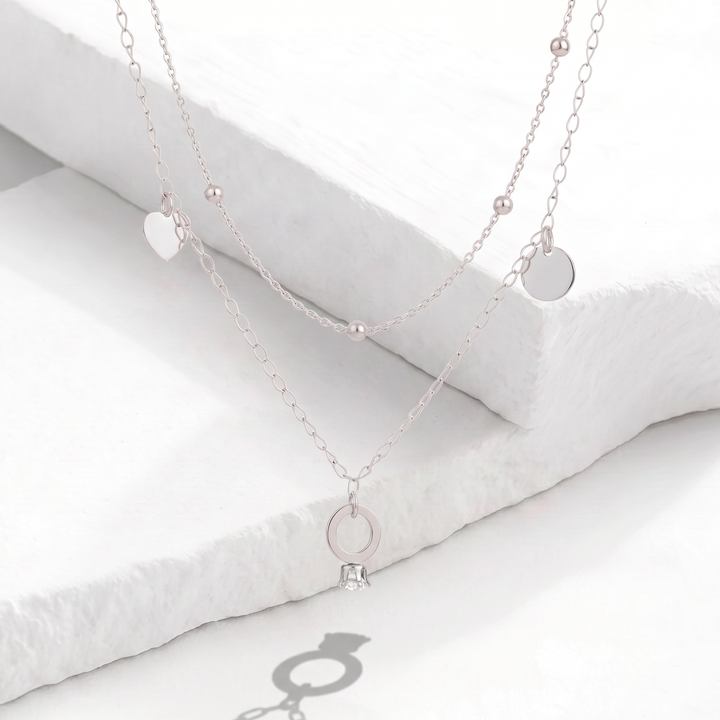 Collar doble cadena para mujer en acero inoxidable plateado con esferas intercaladas y dijes colgantes de corazón, círculo liso y anillo con cristal blanco.