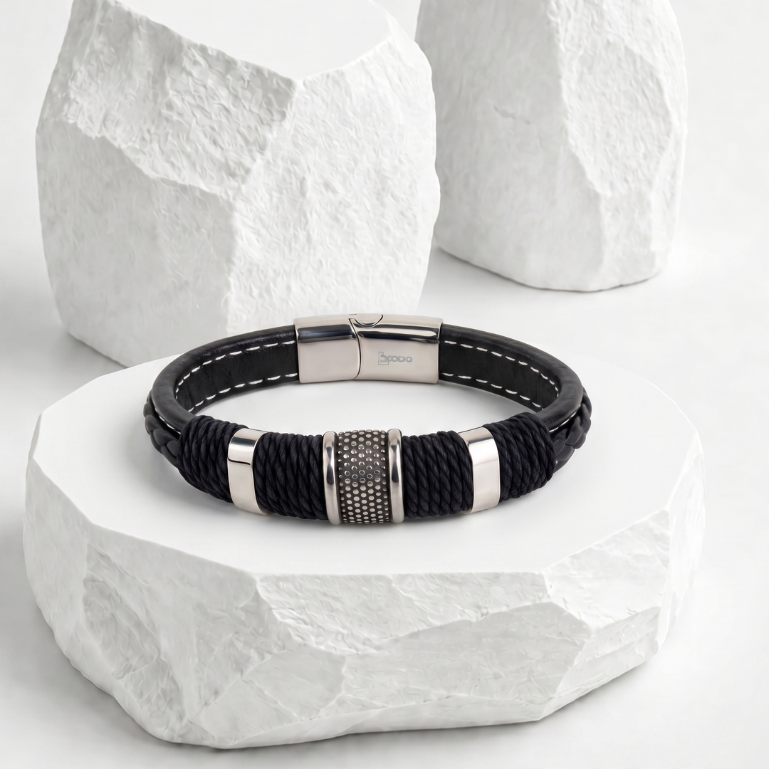 Pulsera para hombre de biocuero negro con costuras blancas, detallada con cordón trenzado negro, pasador central plateado texturizado y broche rectangular magnético de acero inoxidable.
