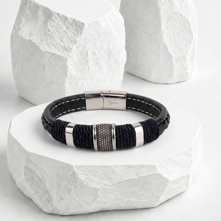Pulsera para hombre de biocuero negro con costuras blancas, detallada con cordón trenzado negro, pasador central plateado texturizado y broche rectangular magnético de acero inoxidable.