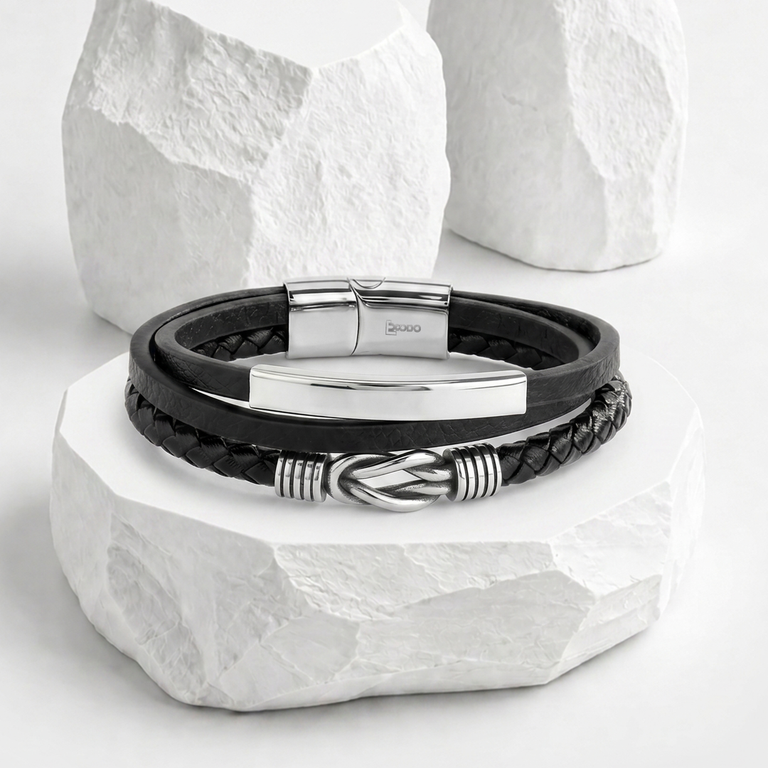 Pulsera para hombre marca Bodo de biocuero negro con diseño de triple capa, equipada con una placa horizontal lisa plateada, dije de nudo marino y broche magnético de acero inoxidable.