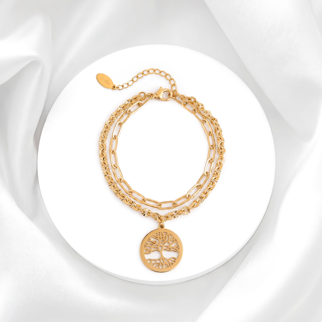 Pulsera dorada de mujer en acero 316L con baño en oro de 18k, diseño de doble cadena y dije circular de Árbol de la Vida sobre soporte blanco.