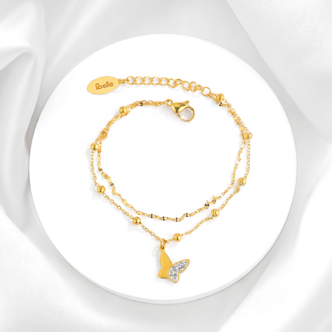Pulsera de doble cadena para mujer en acero quirúrgico 316 con baño de oro de 18k, decorada con esferas doradas lisas y un dije de mariposa de perfil con cristales blancos.