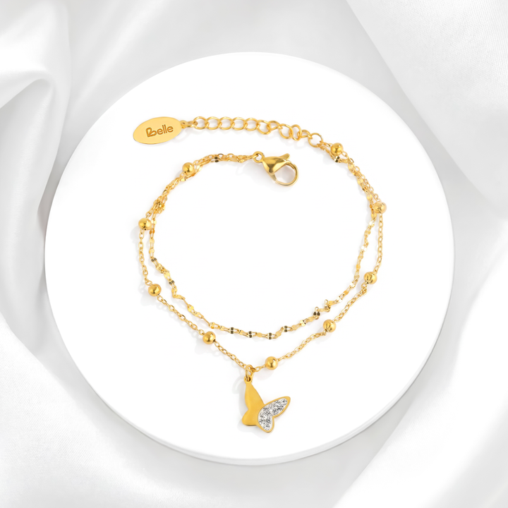 Pulsera de doble cadena para mujer en acero quirúrgico 316 con baño de oro de 18k, decorada con esferas doradas lisas y un dije de mariposa de perfil con cristales blancos.