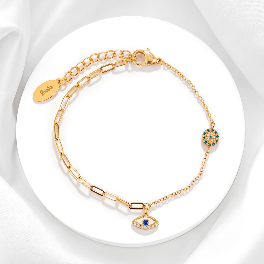 Pulsera mística de mujer en acero quirúrgico 316L baño oro 18k con cadena híbrida de eslabones y dijes de ojo turco con cristales azules y blancos sobre soporte circular.