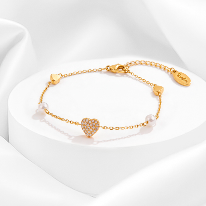 Pulsera fina de mujer en acero quirúrgico 316 baño oro 18 quilates con dije central de corazón con cristales blancos y detalles de perlas sobre soporte circular.