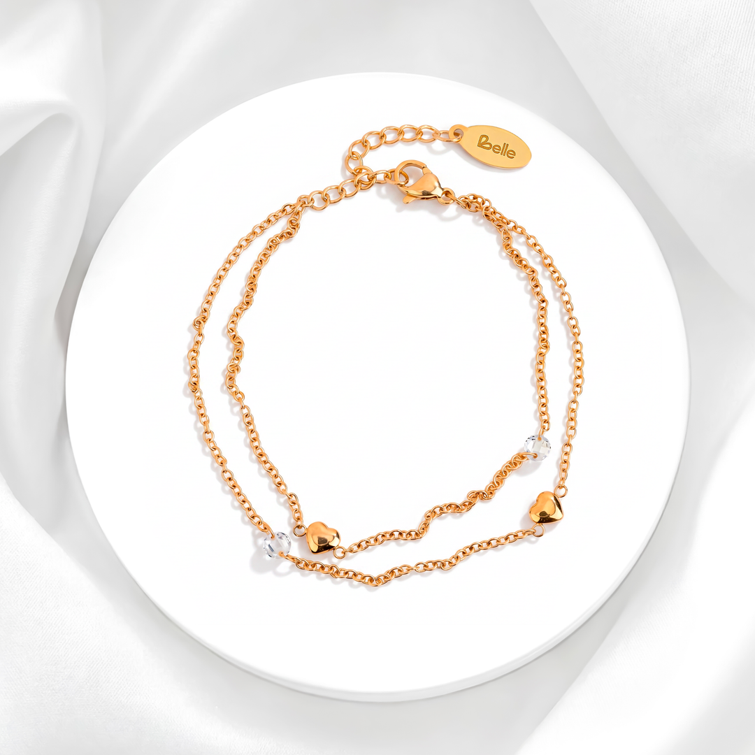 Pulsera dorada de mujer en acero quirúrgico 316 baño oro de 18 quilates con diseño de doble cadena, dijes de corazones lisos pulidos y cristales blancos sobre base circular blanca.