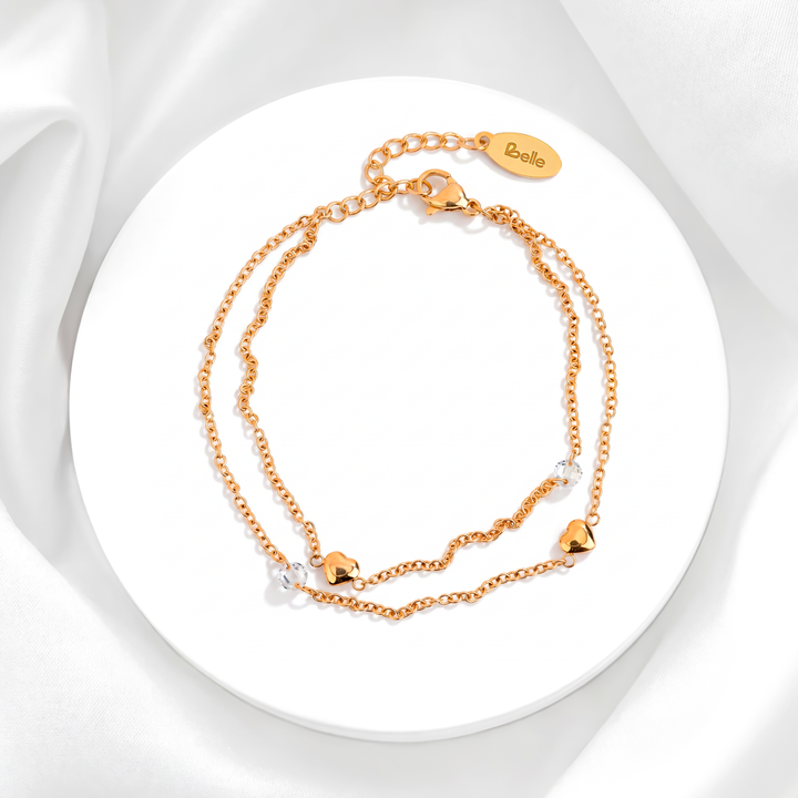 Pulsera dorada de mujer en acero quirúrgico 316 baño oro de 18 quilates con diseño de doble cadena, dijes de corazones lisos pulidos y cristales blancos sobre base circular blanca.