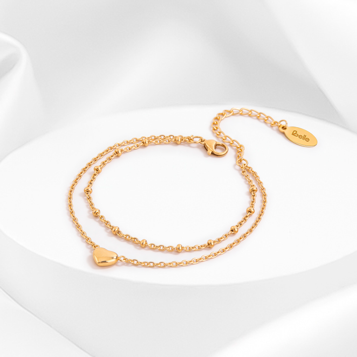 Pulsera dorada de mujer en acero quirúrgico 316 baño oro de 18 quilates con diseño de doble cadena, dijes de corazones lisos pulidos y cristales blancos sobre base circular blanca.