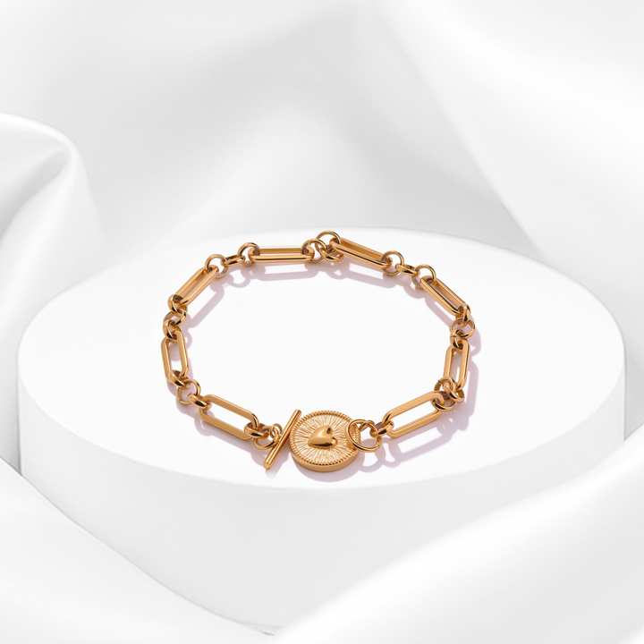Pulsera dorada de eslabones para mujer en acero quirúrgico 316 baño oro 18 quilates con dije circular de medallón con corazón labrado vintage sobre base blanca.