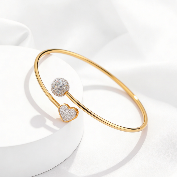Brazalete rígido abierto de mujer en acero quirúrgico 316 baño oro 18 quilates con dijes frontales de esfera redonda y corazón con pavé de cristales blancos sobre base circular.
