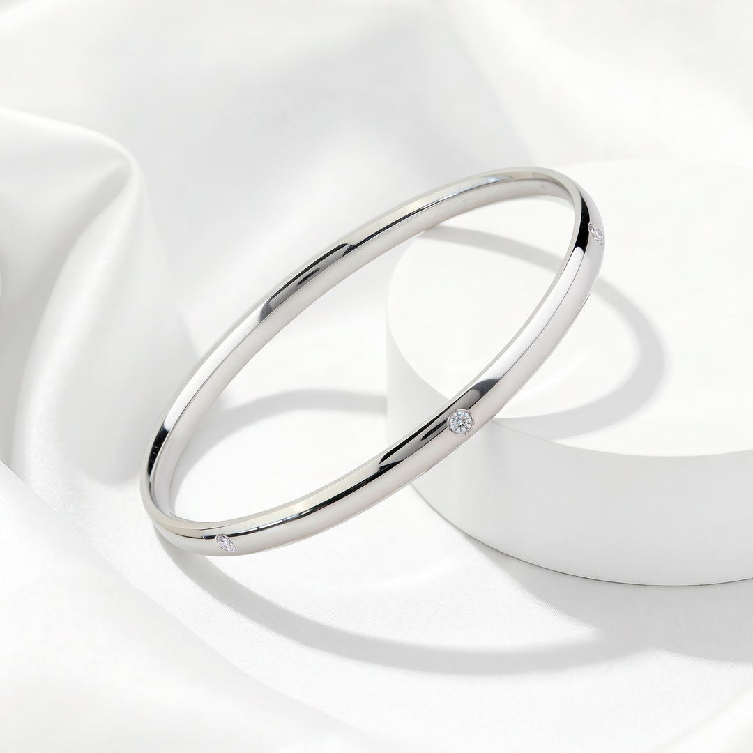 Brazalete rígido de mujer en acero 316L plateado con pequeños cristales blancos incrustados sobre soporte circular blanco.