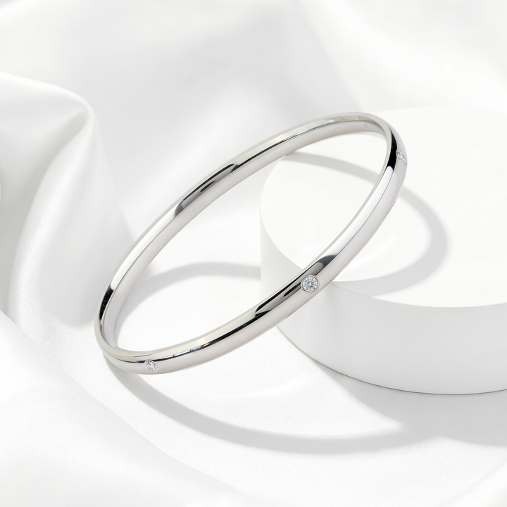 Brazalete rígido de mujer en acero 316L plateado con pequeños cristales blancos incrustados sobre soporte circular blanco.