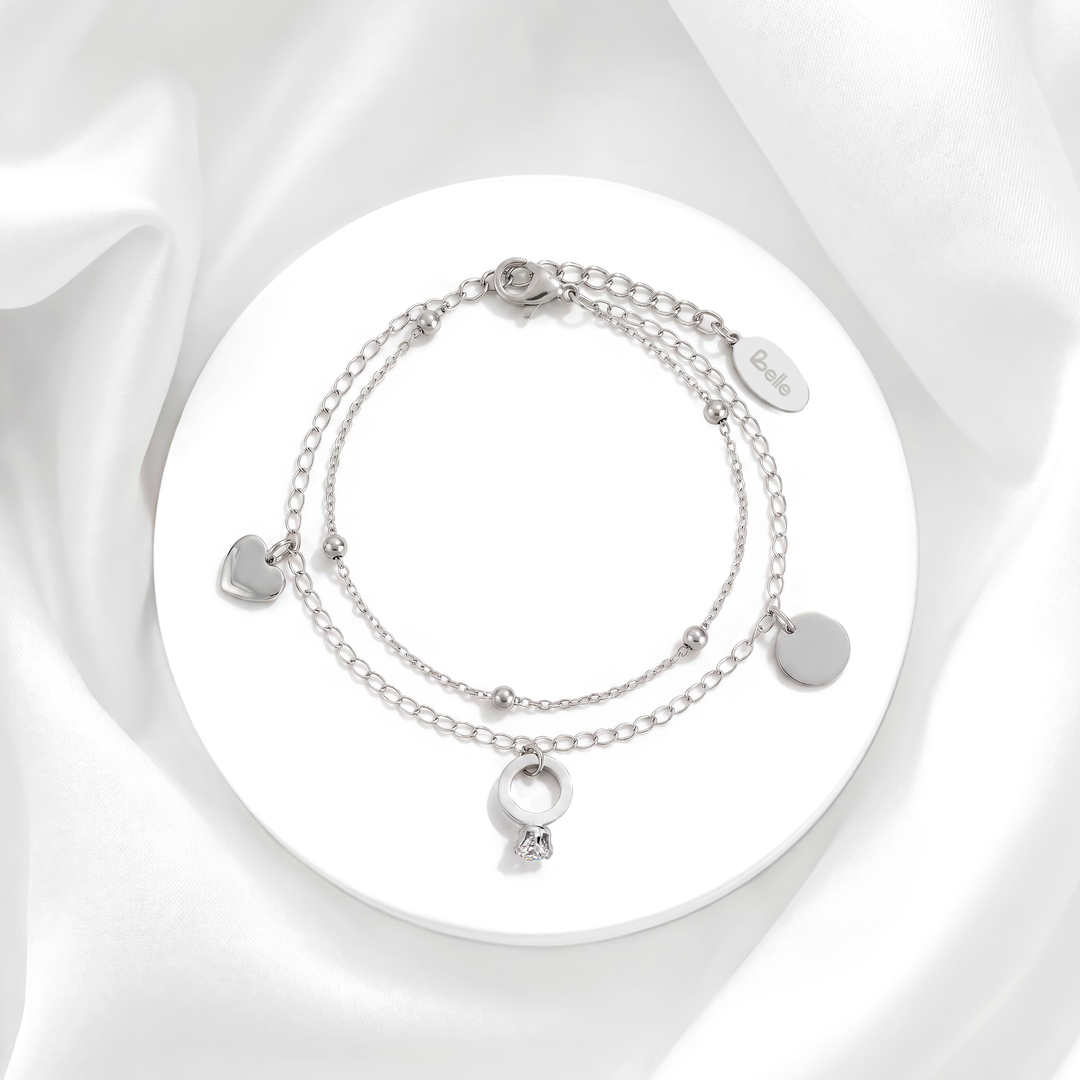 Pulsera plateada de mujer de doble cadena en acero quirúrgico 316 con dijes entrelazados de corazón liso, anillo circular y un cristal blanco sobre soporte circular.