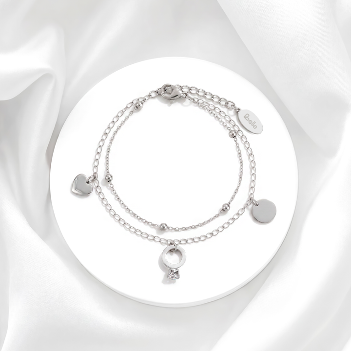 Pulsera plateada de mujer de doble cadena en acero quirúrgico 316 con dijes entrelazados de corazón liso, anillo circular y un cristal blanco sobre soporte circular.