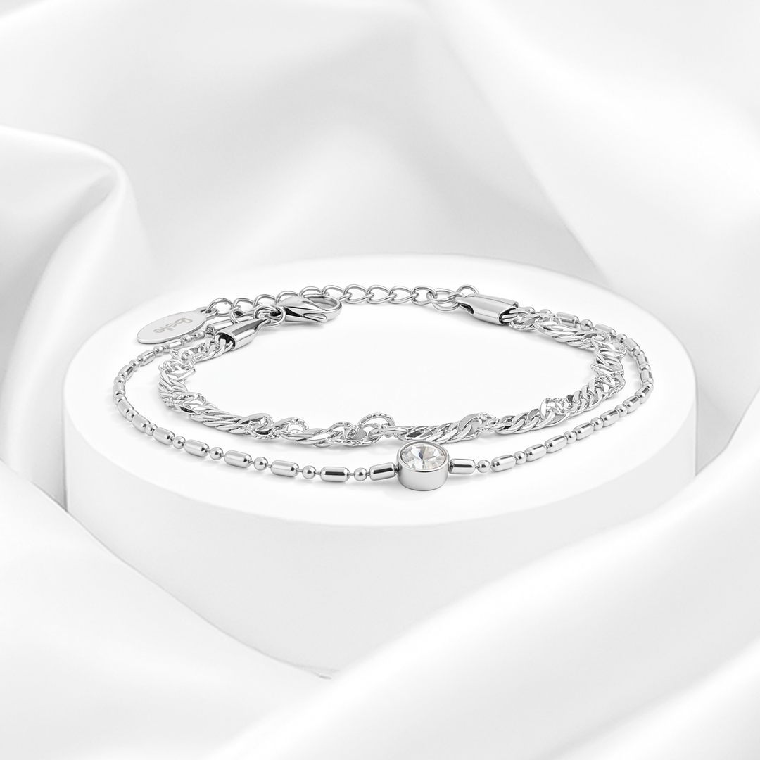 Pulsera plateada de mujer de doble cadena en acero quirúrgico 316 con un hilo trenzado y otro de eslabones con dije circular de cristal blanco solitario sobre expositor redondo.