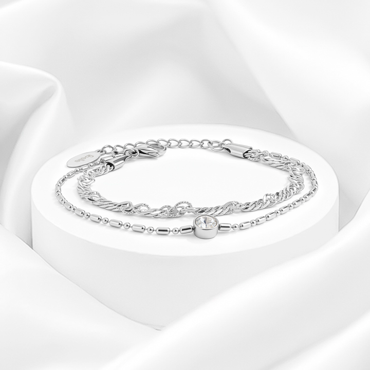 Pulsera plateada de mujer de doble cadena en acero quirúrgico 316 con un hilo trenzado y otro de eslabones con dije circular de cristal blanco solitario sobre expositor redondo.