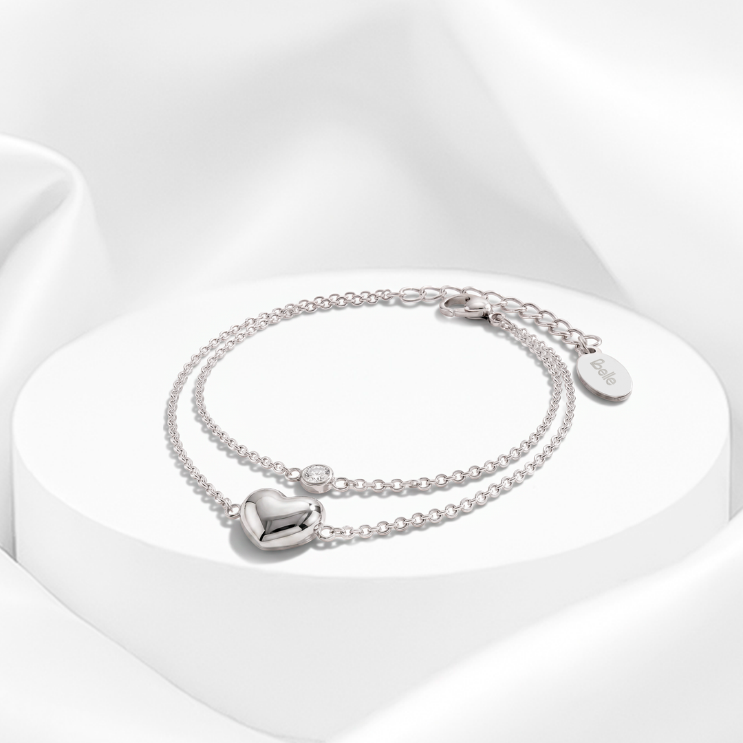 Pulsera plateada de doble cadena fina para mujer en acero quirúrgico 316 con un dije de corazón abombado pulido y un cristal blanco solitario sobre superficie redonda.