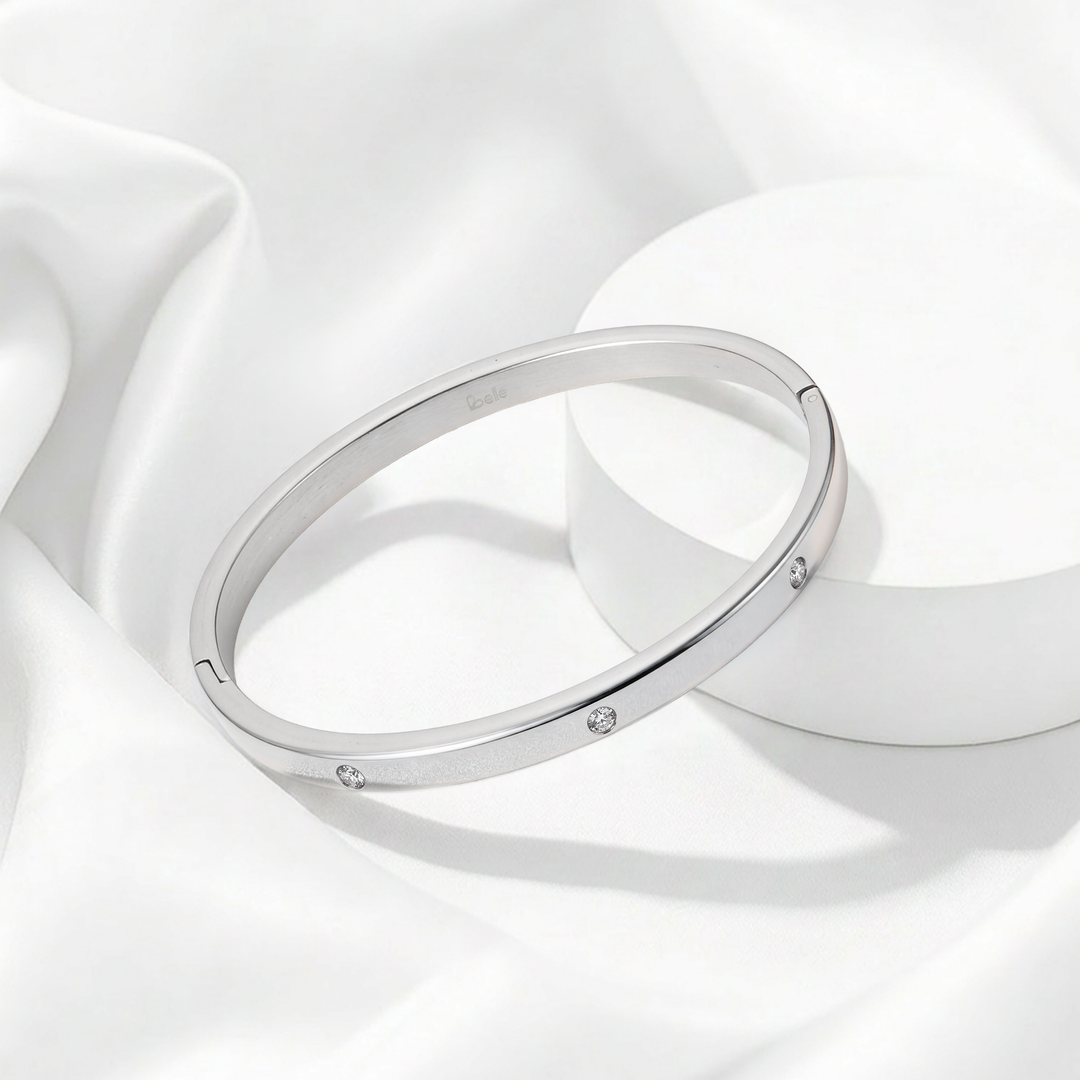 Brazalete rígido de mujer de perfil plano en acero quirúrgico 316 plateado, con superficie lisa pulida a espejo e incrustaciones de cristales blancos circulares.