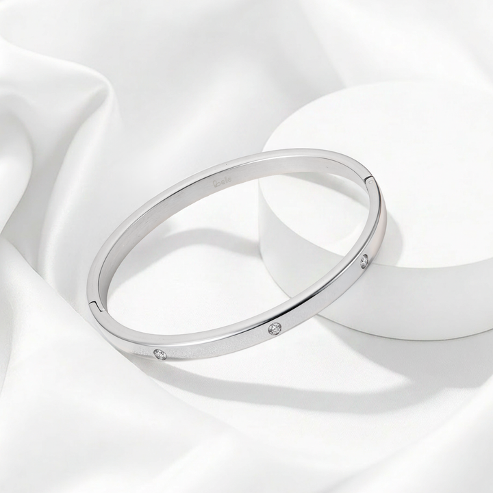 Brazalete rígido de mujer de perfil plano en acero quirúrgico 316 plateado, con superficie lisa pulida a espejo e incrustaciones de cristales blancos circulares.